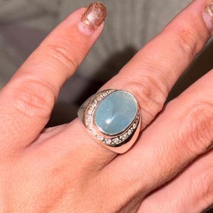 Op de afbeelding: Een zilveren ring met een grote, ovale, lichtblauwe edelsteen. De edelsteen is omgeven door een rij kleinere, heldere stenen. De ring wordt aan een vinger gedragen, waarbij het ontwerp en de kleur van het sieraad worden getoond.