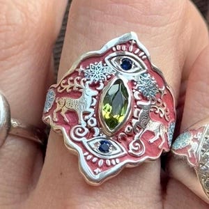 May include: A silver ring with a red enamel background featuring deer, snowflakes, and eye designs. The ring has a central green gemstone and two blue gemstones. A unique piece of jewellery.