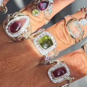 May include: A collection of silver rings featuring various gemstones including a pear-shaped deep red stone, a square-cut green stone, a rectangular dark red stone, and an iridescent opal, all accented with small clear stones in ornate settings.