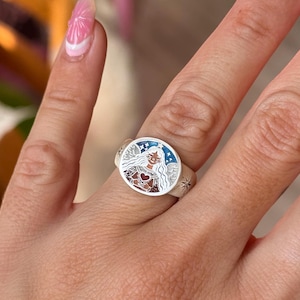 Puede incluir: Anillo de plata con una cara redonda que presenta una ilustraci&oacute;n detallada de una mujer con alas de &aacute;ngel, un coraz&oacute;n y estrellas. El anillo tiene un borde azul y blanco y se lleva en un dedo.