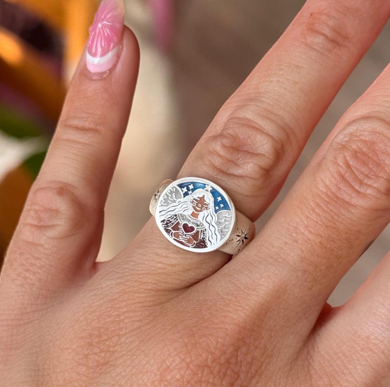 Puede incluir: Anillo de plata con una cara redonda que presenta una ilustraci&oacute;n detallada de una mujer con alas de &aacute;ngel, un coraz&oacute;n y estrellas. El anillo tiene un borde azul y blanco y se lleva en un dedo.