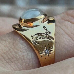 May include: A gold-colored ring featuring a large, round, light-colored stone. The ring has a detailed design of a rabbit and stars with a small diamond accent. The ring is held by a person.