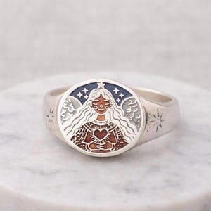 Puede incluir: Un anillo de plata con una cara ovalada que presenta una ilustraci&oacute;n detallada. La ilustraci&oacute;n representa a una mujer con pelo largo y blanco, sosteniendo un coraz&oacute;n, rodeada de estrellas y alas. El anillo tiene dise&ntilde;os de estrellas en los lados.