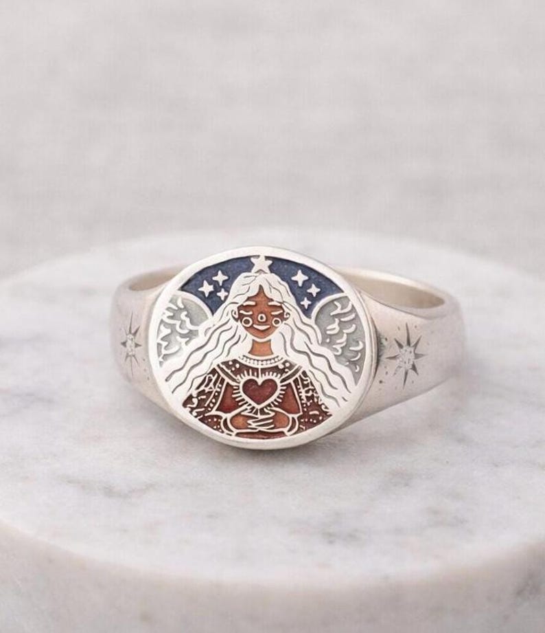 Puede incluir: Un anillo de plata con una cara ovalada que presenta una ilustraci&oacute;n detallada. La ilustraci&oacute;n representa a una mujer con pelo largo y blanco, sosteniendo un coraz&oacute;n, rodeada de estrellas y alas. El anillo tiene dise&ntilde;os de estrellas en los lados.