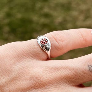 May include: A silver ring featuring a mushroom design with red and white enamel details. The ring has a detailed, nature-inspired design, with a mushroom at the center and star-like accents. The ring is worn on a finger.