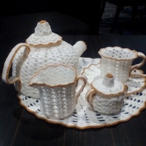 Puede incluir: Un juego de té de ganchillo blanco con una tetera, una jarra de crema, un azucarero y una taza y un platillo. El juego está sobre un encaje de ganchillo blanco.