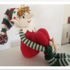 May include: A knitted elf doll with a red and white striped hat, green and white striped sleeves, and red trousers. The elf is sitting with its legs crossed and its arms crossed.