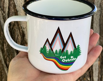 May include: White enamel mug with a black rim and handle. The mug features a mountain graphic with a rainbow and the text "Get Outside". The mug is held in a hand.