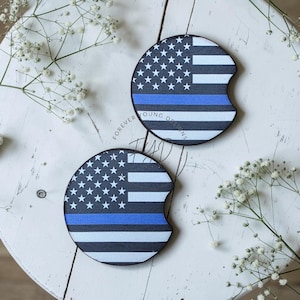 May include: Two circular car cup holder coasters featuring a graphic of the American flag with a horizontal blue line across the center. Each coaster has a small semi-circular notch on the edge. The text Forever Young Designs is visible.