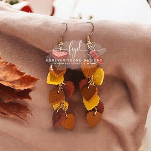 May include: A pair of dangle earrings with a cascade of layered fall leaves in shades of brown, orange, yellow, and pink. The earrings are made of polymer clay and have gold-toned hooks. The text "Forever Young Designs Handmade Crafts & More" is visible in the image.