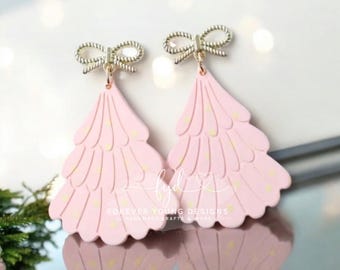 Può includere: Un paio di orecchini pendenti rosa chiaro a forma di alberi di Natale stilizzati con bordi smerlati e piccoli punti gialli. Ogni orecchino presenta un fiocco in metallo dorato sulla parte superiore. Il testo Forever Young Designs è visibile.