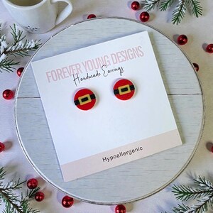 May include: A pair of red and black Santa belt earrings on a white card. The card reads "FOREVER YOUNG DESIGNS Handmade Earrings" and "Hypoallergenic". The earrings are round with a red background, black belt, and gold buckle.