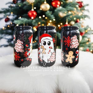 May include: Three black glass tumblers with lids, each featuring a white ghost wearing a red Santa hat, gingerbread cookies, candy canes, and red snowflake patterns. The text FOREVER YOUNG DESIGNS HANDMADE CRAFTS & MORE is printed at the bottom.