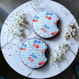 May include: Two circular car cup holder coasters featuring a light blue and white horizontal striped pattern with red cherry illustrations, blue bows, and small blue stars. The text Forever Young Designs is printed on the surface.