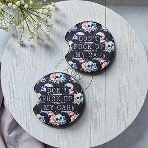 May include: Two circular car cup holder coasters featuring a black background with a repeating pattern of skulls and pink flowers. The text DON'T FUCK UP MY CAR is printed in white capital letters across the center of each coaster.