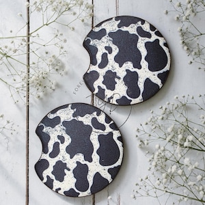 May include: Two circular car coasters featuring a black and white cow print pattern. Each coaster has a small semi-circular notch cut out from the side. The surface of the coasters appears textured with a repeating organic spot design.