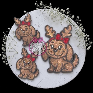 May include: Three glittery brown dog-shaped ornaments with reindeer antlers and red bows. The dogs have black outlines and smiling expressions. A circular logo in the center reads Forever Young Designs 919 with a checkered pattern.