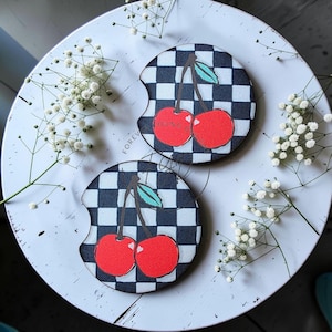 May include: Two circular car cup holder coasters featuring a black and white checkered pattern. Each coaster displays a graphic of two bright red cherries with green leaves attached to brown stems. The design includes small white heart accents.