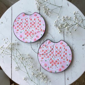 May include: Two circular car cup holder coasters featuring a pink background with a repeating strawberry and white flower pattern. The word MAMA is printed in large white block letters with a red strawberry fill, accompanied by a red bow graphic.