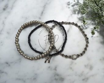 Può includere: Un set di tre braccialetti di perline con perline argentate metallizzate, piccole perline nere e perline scure sfaccettate. Un braccialetto include un ciondolo a forma di fiocco argentato e un altro presenta una perlina rotonda incastonata con cristalli chiari. Il testo Forever Designs F&D è visibile.