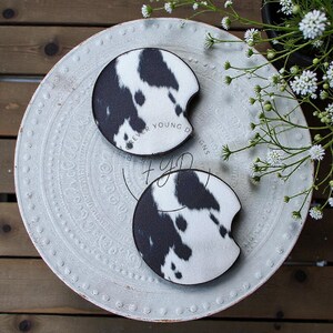 May include: Two circular car cup holder coasters featuring a black and white cow print pattern. Each coaster has a small semi-circular notch on the side. The text Forever Young Designs and the initials FYD are printed on the surface of the coasters.