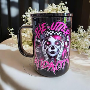 May include: Black mug with a handle and lid, featuring a graphic of a woman with pink hair and a skull face. The text "THE UTTER AUDACITY" is displayed in pink. The mug is on a white surface.