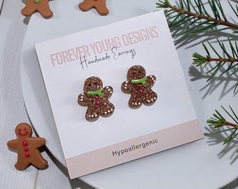 Può includere: Un paio di orecchini a lobo a forma di omino di pan di zenzero marrone glitterato su un cartoncino bianco con la scritta Forever Young Designs Handmade Earrings e Hypoallergenic, con sciarpe verdi e bottoni rossi.