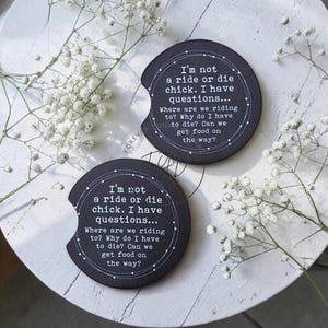 May include: Two round black car cup holder coasters featuring a white circular border and the text: I'm not a ride or die chick. I have questions... Where are we riding to? Why do I have to die? Can we get food on the way? Each coaster has a notch.