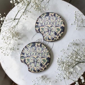 May include: Two circular car cup holder coasters featuring a camouflage pattern in shades of green, brown, and tan. Each coaster displays the word MAMA in a bold, block font above a graphic illustration of a large, decorative hair bow.