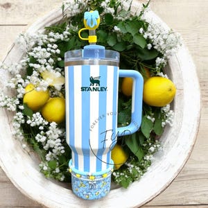 May include: A blue and white striped stainless steel travel mug with a handle and a clear lid. The mug has a blue silicone straw and a blue and white silicone straw topper with a yellow bow. The mug is sitting in a white bowl with white flowers and lemons.