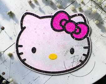 May include: A glittery Hello Kitty character cutout featuring a pale pink face, a vibrant hot pink bow, black glittery whiskers, black eyes, and a yellow nose. The design is outlined in black glitter and includes the text Forever Young Designs.