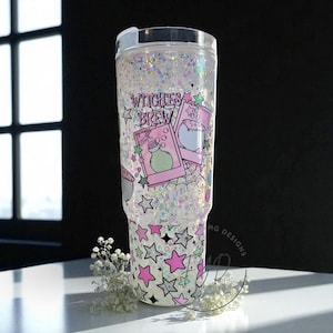 May include: A clear tumbler filled with iridescent glitter and a decorative design. The design includes the text "Witches Brew", pink stars, and illustrations of potion bottles and a cauldron. The bottom of the tumbler features a pattern of pink and black stars.