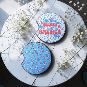 May include: Two circular car cup holder coasters with a notch. One features a blue floral pattern with red and blue stars and the text She loves JESUS and AMERICA too. The other has a light blue background with a repeating pattern of red and white stars.