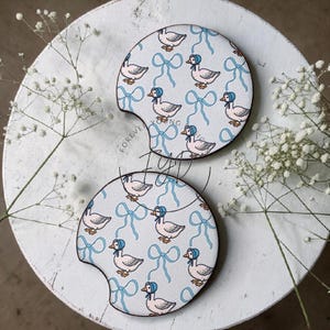May include: Two round car cup holder coasters featuring a repeating pattern of white ducks wearing blue bonnets and light blue ribbons on a white background. Each coaster has a small semi-circular notch on the side for easy removal from the holder.