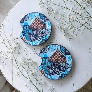 May include: Two circular car cup holder coasters featuring a blue and white distressed background with a brown checkered spade design, blue lace-like floral accents, and the text a lil bougie written in a black script font across the center.