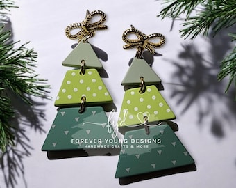 Può includere: Un paio di orecchini pendenti con un fiocco dorato in alto e tre segmenti trapezoidali impilati in tonalità di verde. I segmenti presentano pois e motivi a triangolo bianchi. Il testo recita: fyd FOREVER YOUNG DESIGNS HANDMADE CRAFTS & MORE.