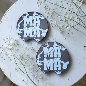May include: Two circular car cup holder coasters featuring a black and white cow print pattern. The word MAMA is printed in large, bold, light-colored block letters in the center of each coaster. Each coaster has a small semi-circular notch at the bottom.