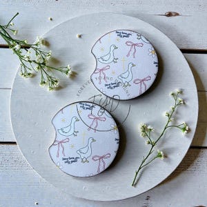 May include: Two circular white car coasters featuring a repeating pattern of geese, pink bows, yellow daisies, and small crosses. The text "Jesus loves you silly goose!" is printed in black script across the surface of each coaster.
