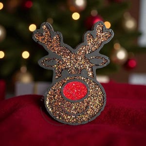 May include: A glittery reindeer decoration with a red nose and antlers. The reindeer is black with gold glitter and a red glitter nose. The background is a blurred Christmas tree with ornaments.