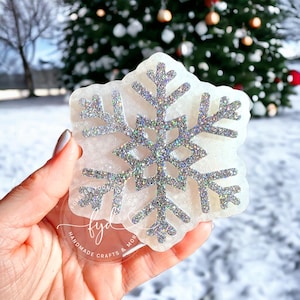 May include: A snowflake-shaped coaster with a glittery silver design on a white background. The coaster is held in a hand, with a blurred winter scene in the background. The coaster is a handmade craft.