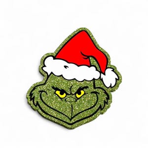 May include: A glittery green character head cutout featuring a mischievous facial expression with yellow eyes and a wide grin. The character wears a bright red Santa hat with a white fluffy trim. The entire piece has a textured, sparkling finish.