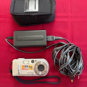May include: A silver Sony Cyber-shot digital camera with a wrist strap, a black Sony power adapter with a long cord, and a black fabric carrying case. The camera features a lens, flash, and a label indicating 4.0 megapixels.