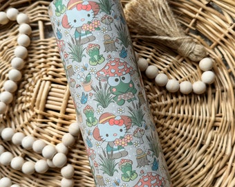 Op de afbeelding: Een cilindervormige beker met een herhalend patroon van stripfiguren, waaronder een witte kat met een rode strik en een groene kikker met een rode paddenstoelhoed, omringd door kleine bloemen, bijen, vlinders en diverse groene planten.