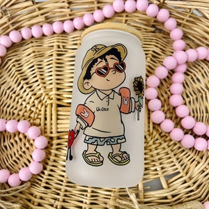 May include: A frosted glass tumbler with a bamboo lid featuring a cartoon character wearing a tan bucket hat, heart-shaped sunglasses, and orange arm floaties. The character holds a sunflower and a red heart icon, with the text Un Coco printed on the shirt.
