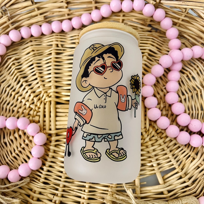 May include: A frosted glass tumbler with a bamboo lid featuring a cartoon character wearing a tan bucket hat, heart-shaped sunglasses, and orange arm floaties. The character holds a sunflower and a red heart icon, with the text Un Coco printed on the shirt.