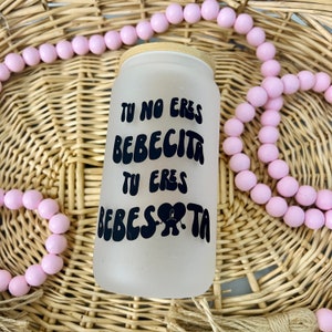 May include: A frosted glass tumbler with a bamboo lid featuring black text that reads TU NO ERES BEBECITA TU ERES BEBESOTA with a heart-shaped graphic incorporating a sad face. The tumbler is surrounded by a string of light pink wooden beads.