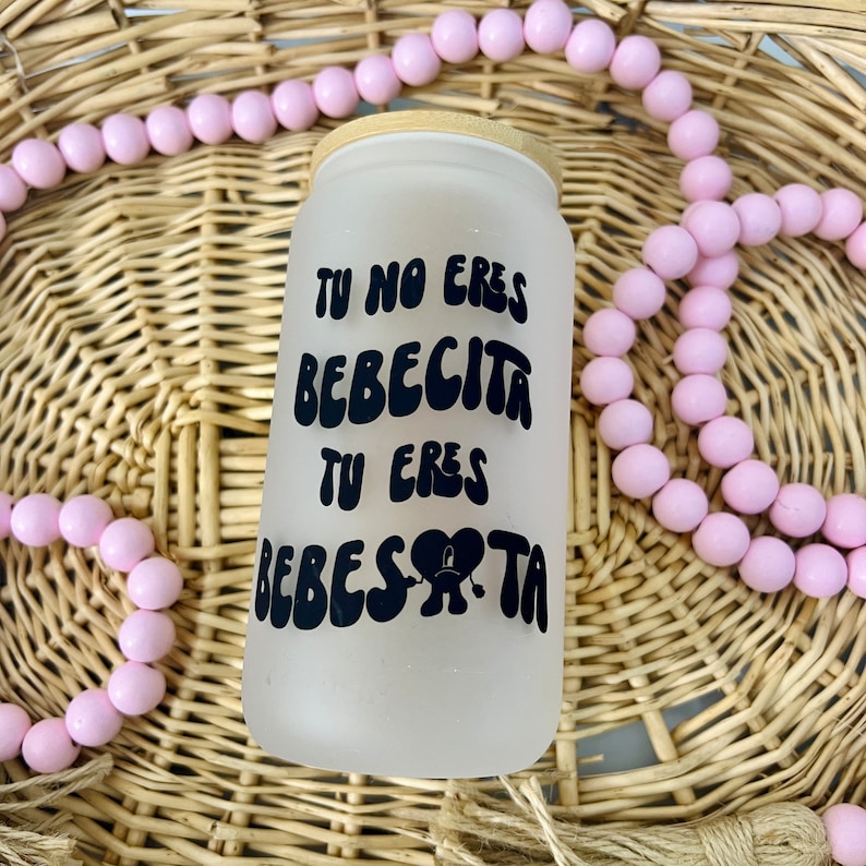 May include: A frosted glass tumbler with a bamboo lid featuring black text that reads TU NO ERES BEBECITA TU ERES BEBESOTA with a heart-shaped graphic incorporating a sad face. The tumbler is surrounded by a string of light pink wooden beads.