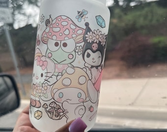 Op de afbeelding: Een matglazen beker met een kleurrijke grafische print van diverse stripfiguren, waaronder een witte kat met een roze strik, een groene kikker en een zwart personage met een bloemenkrans, omringd door paddenstoelen, bijen en kristallen.