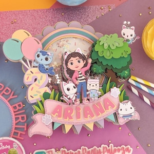 以下が含まれることがあります： ピンクとグリーンの3Dケーキトッパー。ピンクのシャツと白黒のストライプのパンツを着た女の子のキャラクターが描かれています。トッパーには、木、数字の2、名前が「アリアナ」と書かれたバナー、そしていくつかの漫画の猫も含まれています。トッパーはキラキラと輝き、ピンクと白の肉球のデザインが施されています。