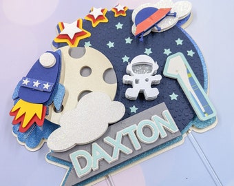 以下が含まれることがあります： 青と銀色のロケット、星、月、宇宙飛行士、そして「DAXTON」という名前が特徴の宇宙をテーマにしたケーキトッパー。1歳の誕生日を祝うことを示唆する数字の「1」も含まれています。デザインにはキラキラとしたアクセントが施されています。